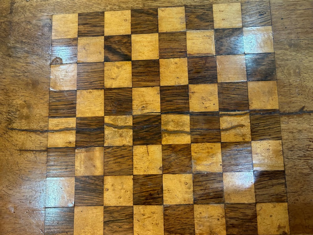 A Victorian warm brown wood games work table featuring an inlaid chessboard top, a single drawer, a damask fabric sewing well, and a turned pedestal base. View VII