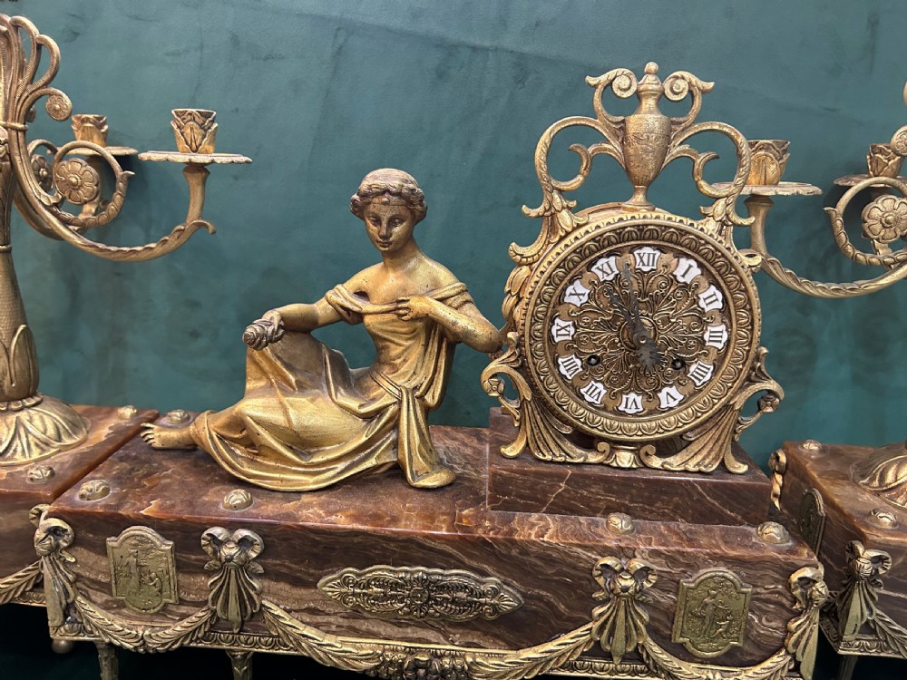 An antique three-piece French ormolu clock garniture featuring a central mantel clock decorated with a classical female figurine and marble base, flanked by two matching five-arm candelabra. View II