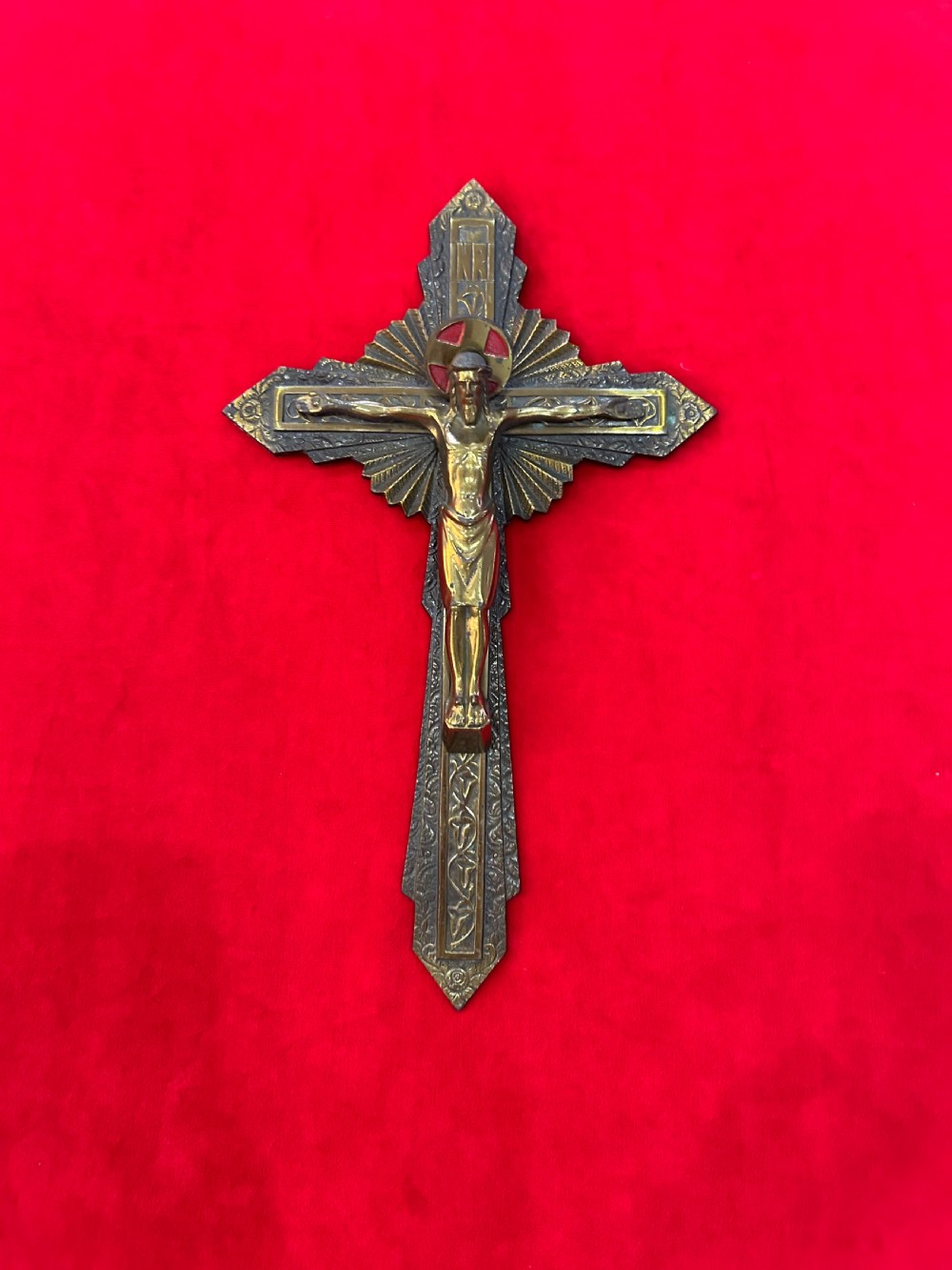 An Art Deco brass crucifix featuring a geometric cross structure with a sunburst halo, red enamelled accents, and a relief figure of Christ.