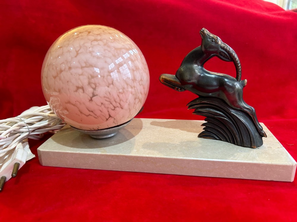 An Art Deco table lamp featuring a rectangular beige marble base, a cast metal leaping gazelle figurine, and a spherical mottled pink glass globe shade.