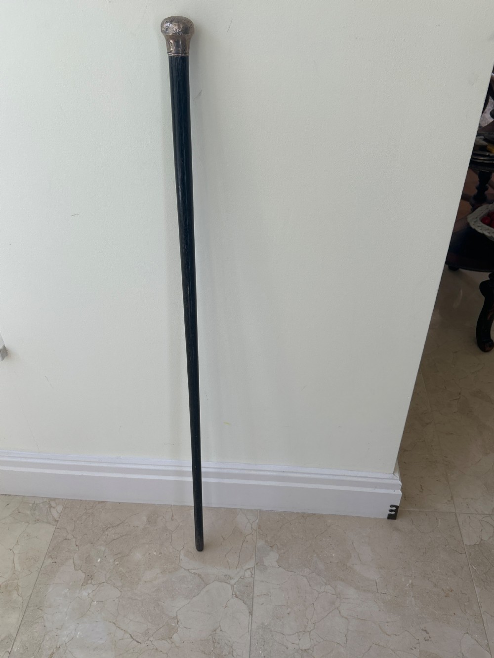 An antique black ebony walking cane featuring a rounded silver top handle standing against a neutral wall.