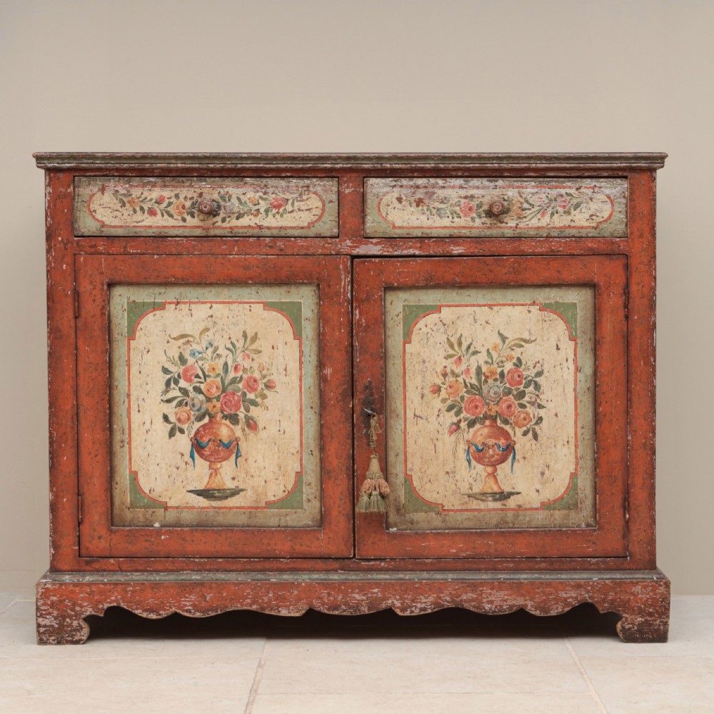 A 19th-century terracotta red painted wooden cabinet featuring two upper drawers and two cupboard doors adorned with cream panels hand-painted with floral urn designs.