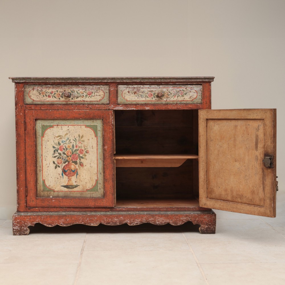 A 19th-century terracotta red painted wooden cabinet featuring two upper drawers and two cupboard doors adorned with cream panels hand-painted with floral urn designs. View IV