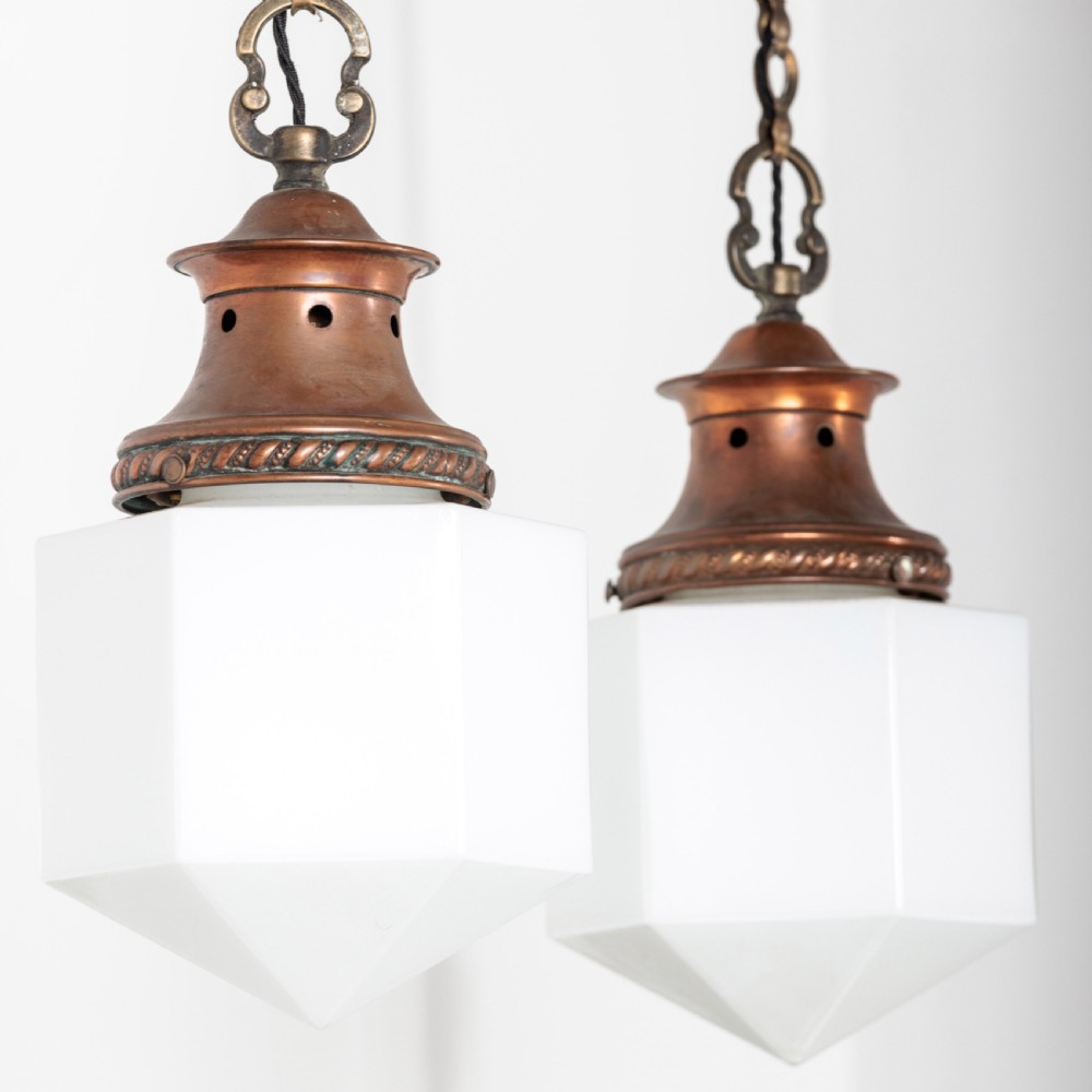 A pair of 1930s Art Deco pendant lights featuring faceted white opal glass hexagonal shades and flared copper top galleries with hanging loop attachments.