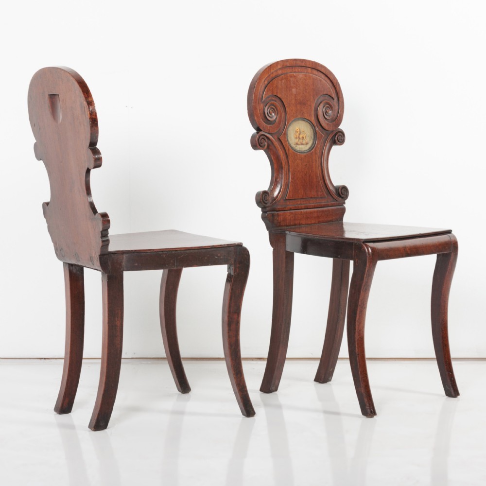 A pair of 19th-century dark mahogany hall chairs featuring scroll-carved backrests with central circular medallions and curved front legs. View X