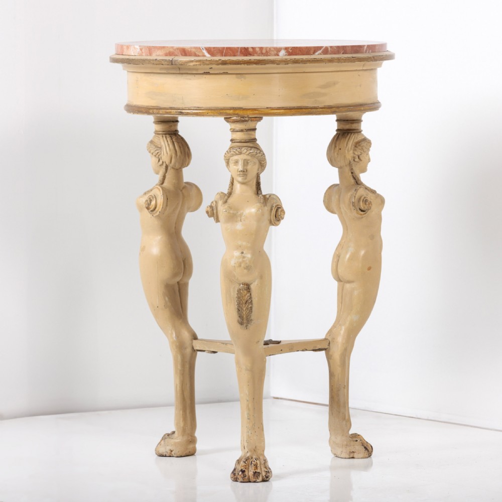 A 19th-century French Empire style circular gueridon table with a pink marble top supported by three carved cream-painted caryatid legs terminating in paw feet.