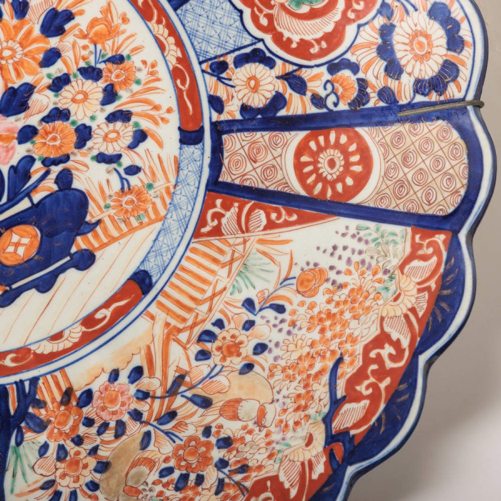 A circular Japanese Imari porcelain charger featuring a scalloped rim, hand-painted cobalt blue and rust red floral motifs, and a central vessel filled with flowers. View IV