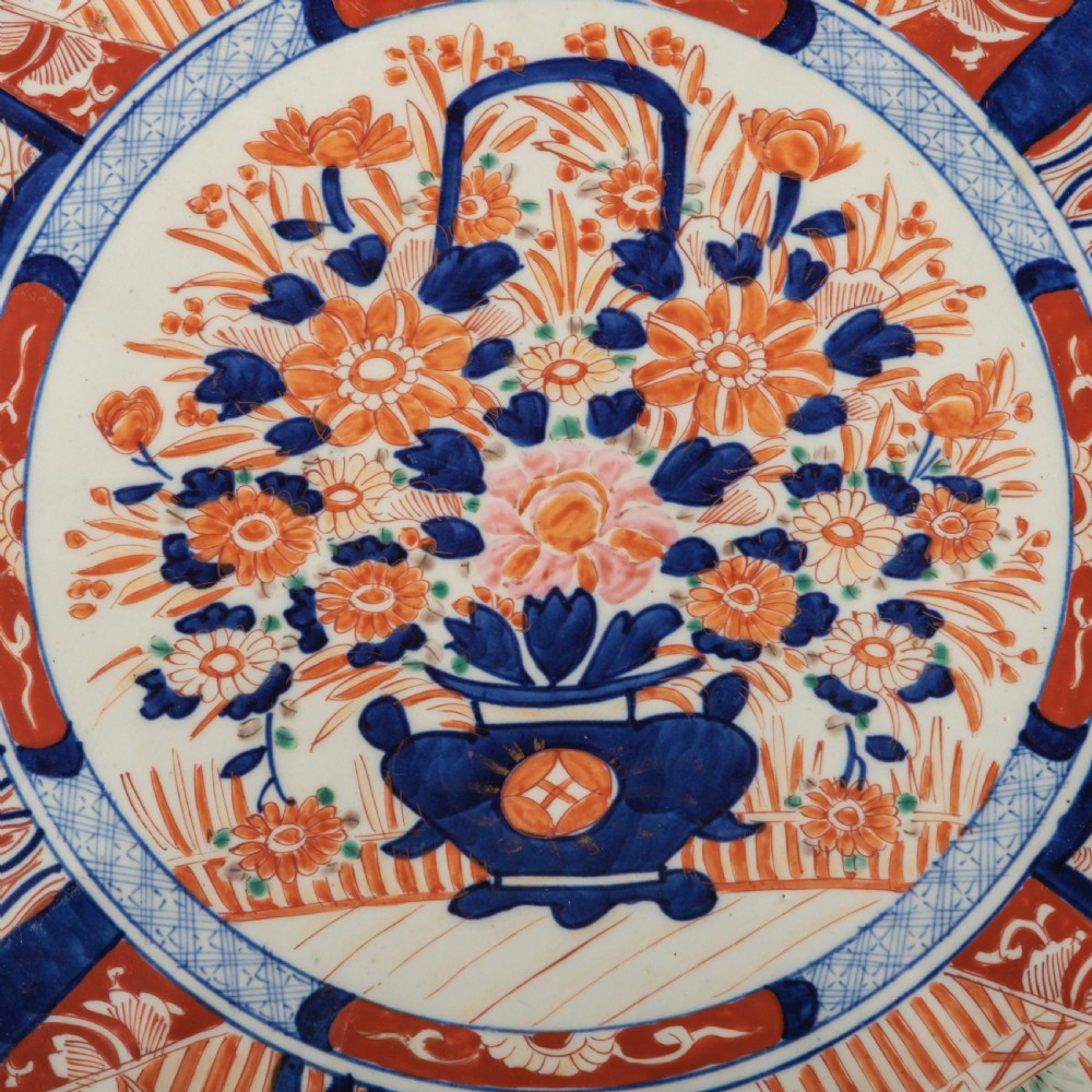 A circular Japanese Imari porcelain charger featuring a scalloped rim, hand-painted cobalt blue and rust red floral motifs, and a central vessel filled with flowers. View V