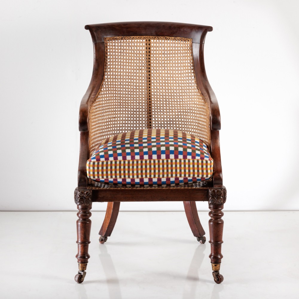 A 19th-century dark timber bergere armchair featuring woven cane back and side panels, turned front legs on brass castors, and a multi-coloured geometric patterned seat cushion.