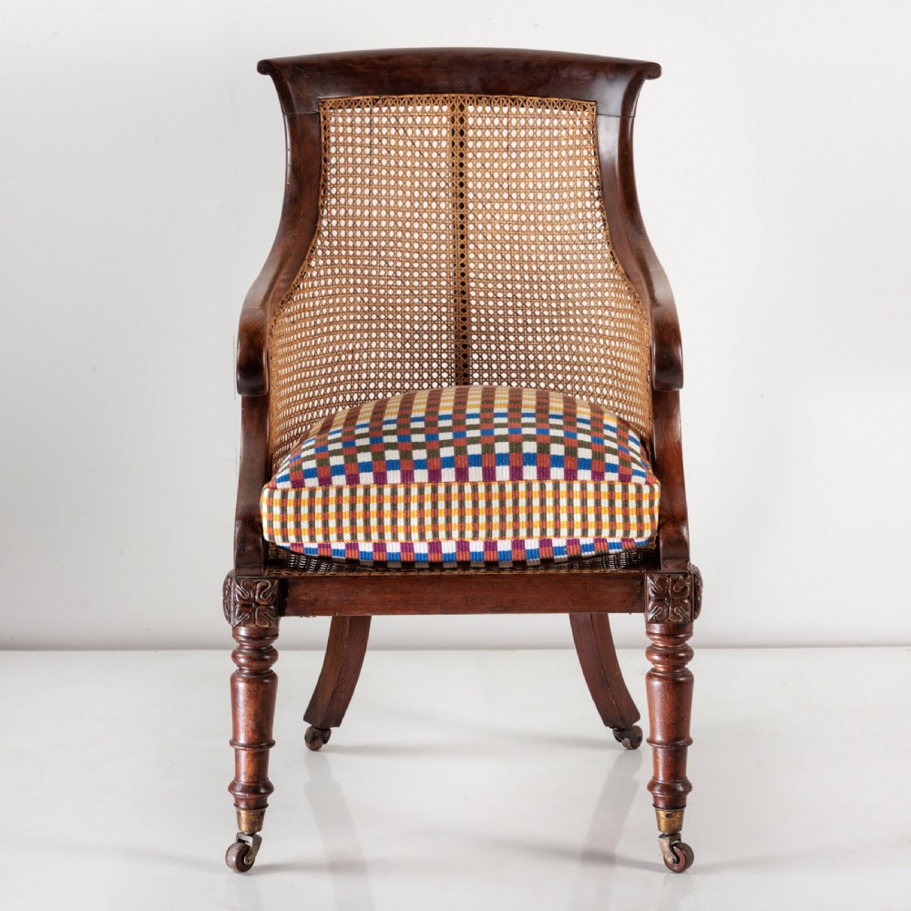 A 19th-century dark timber bergere armchair featuring woven cane back and side panels, turned front legs on brass castors, and a multi-coloured geometric patterned seat cushion. View II