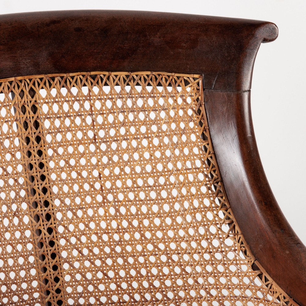 A 19th-century dark timber bergere armchair featuring woven cane back and side panels, turned front legs on brass castors, and a multi-coloured geometric patterned seat cushion. View V