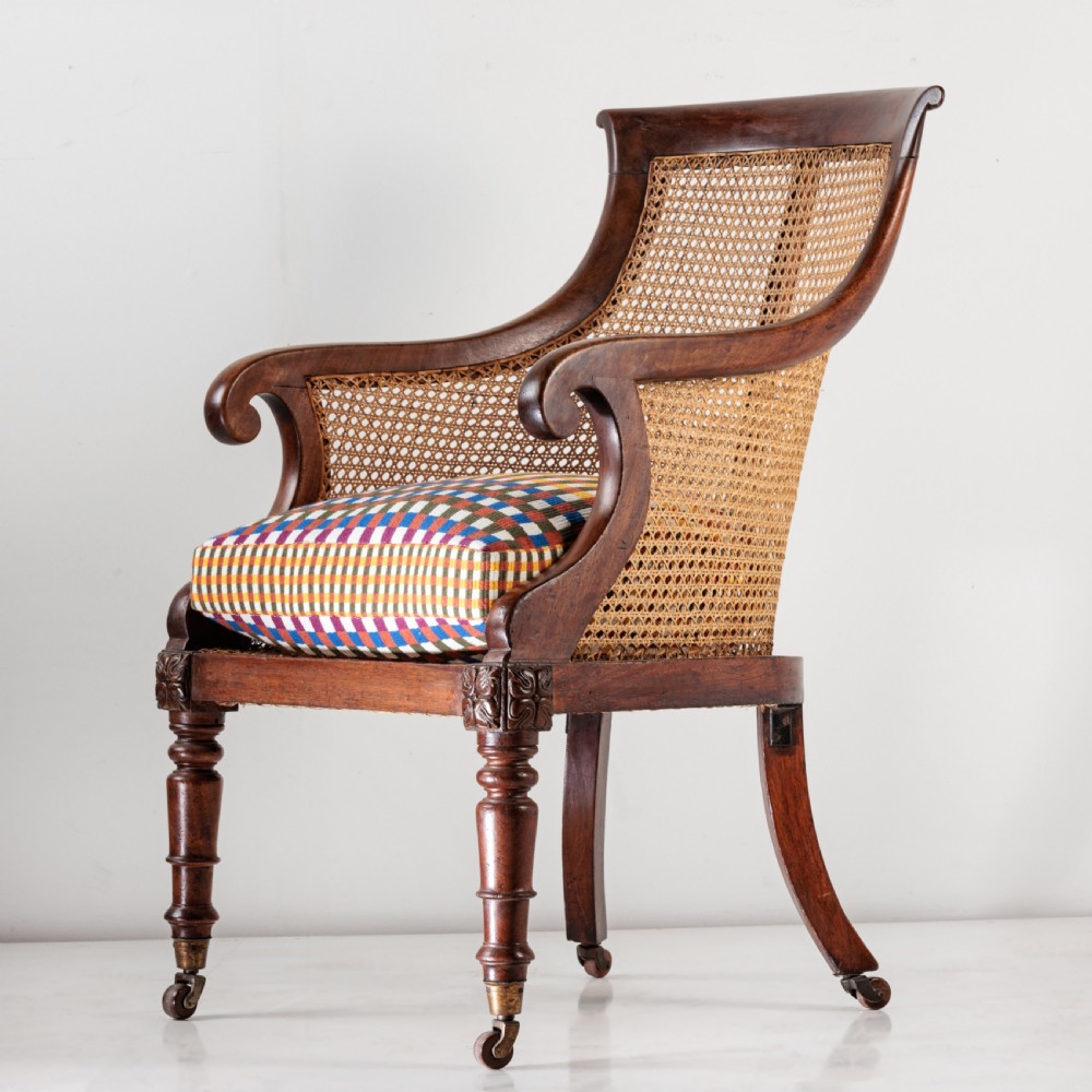 A 19th-century dark timber bergere armchair featuring woven cane back and side panels, turned front legs on brass castors, and a multi-coloured geometric patterned seat cushion. View VI