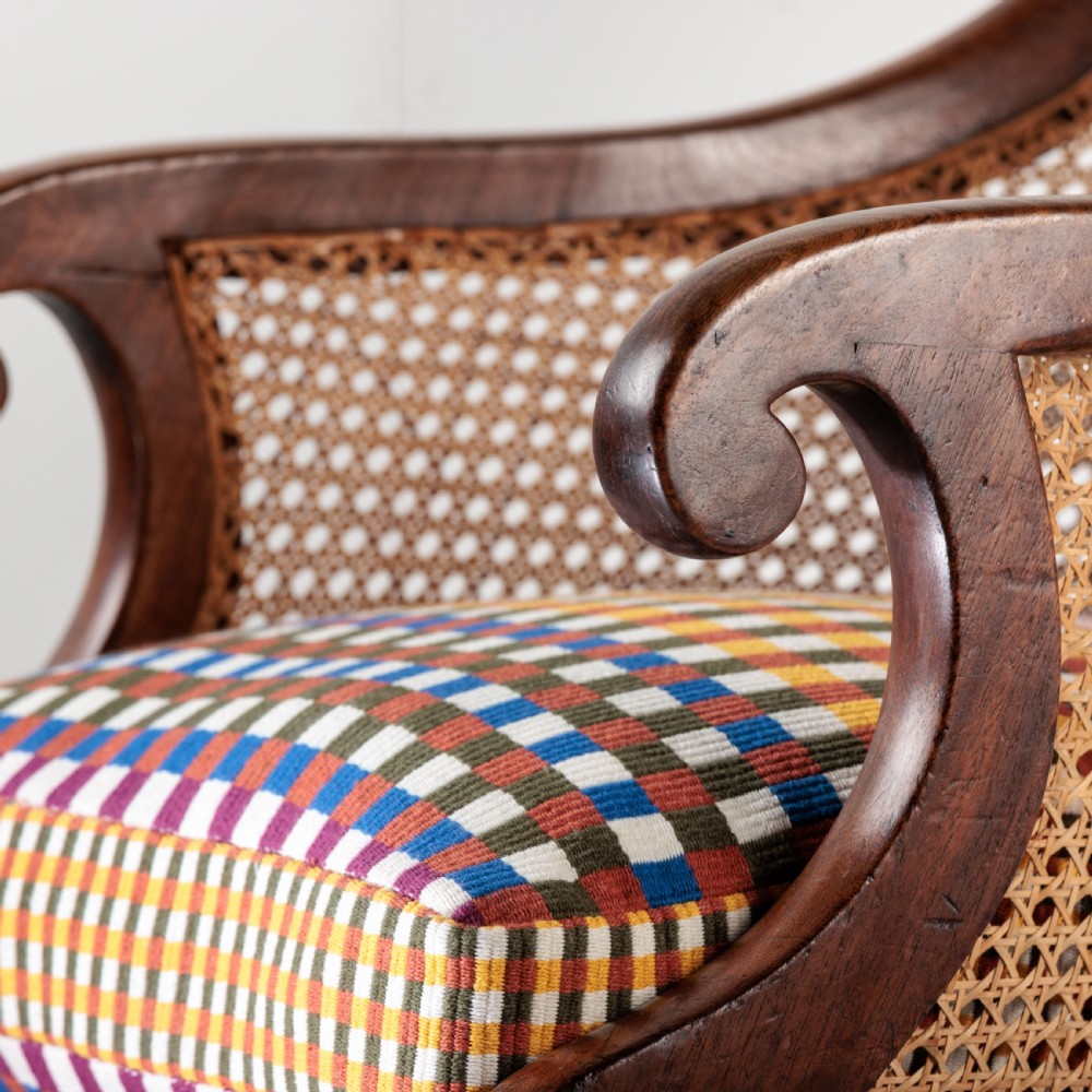 A 19th-century dark timber bergere armchair featuring woven cane back and side panels, turned front legs on brass castors, and a multi-coloured geometric patterned seat cushion. View VII
