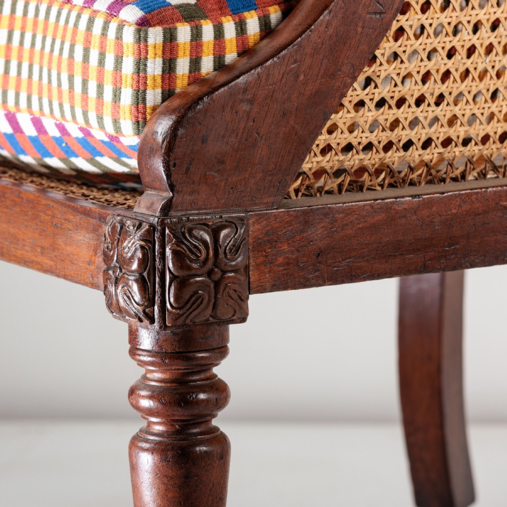 A 19th-century dark timber bergere armchair featuring woven cane back and side panels, turned front legs on brass castors, and a multi-coloured geometric patterned seat cushion. View VIII