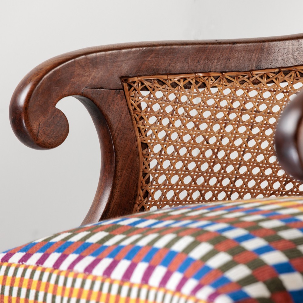 A 19th-century dark timber bergere armchair featuring woven cane back and side panels, turned front legs on brass castors, and a multi-coloured geometric patterned seat cushion. View IX