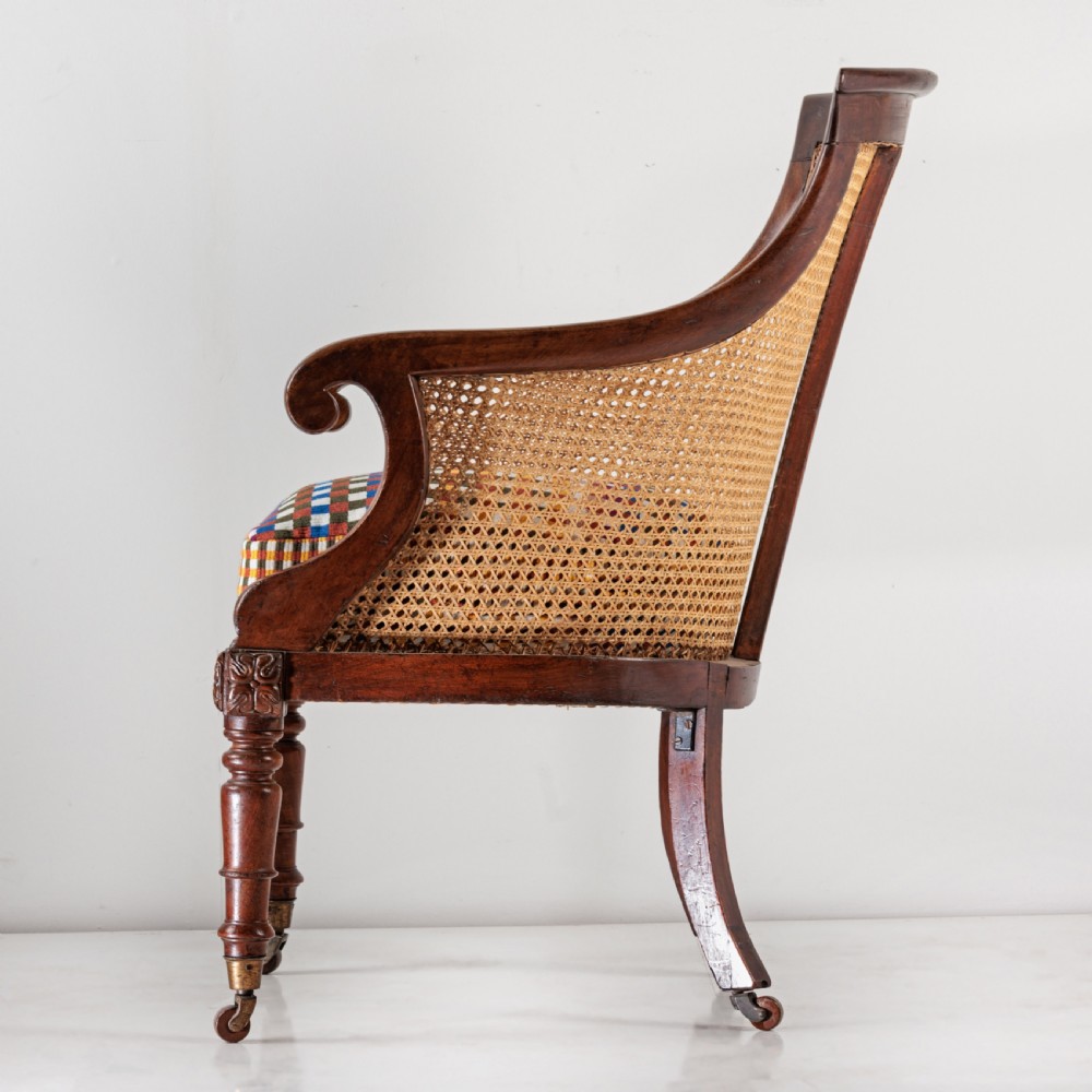 A 19th-century dark timber bergere armchair featuring woven cane back and side panels, turned front legs on brass castors, and a multi-coloured geometric patterned seat cushion. View X