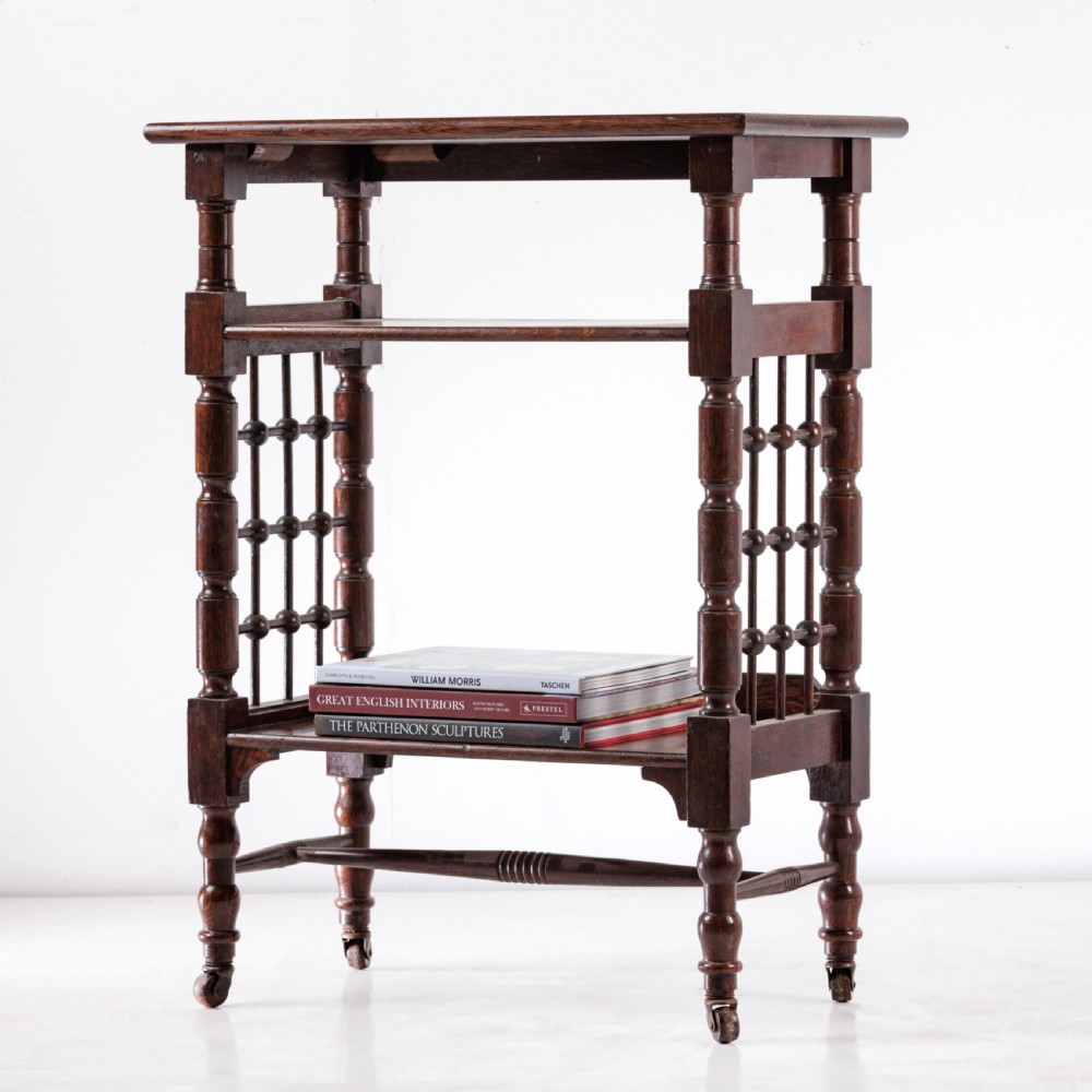 A dark oak Aesthetic Movement side table featuring turned spindle supports, side spindle galleries, three tiers, and legs terminating in castors.