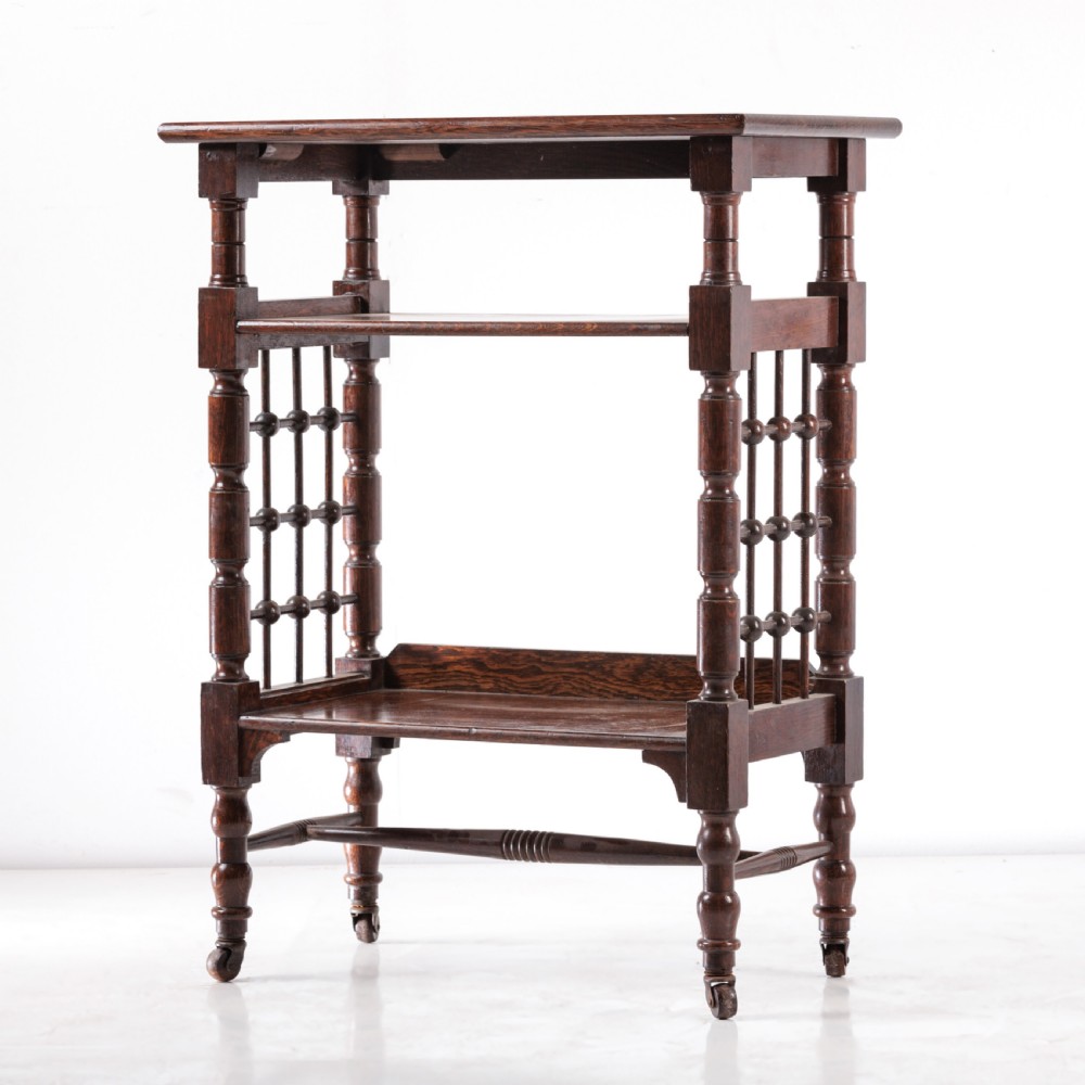 A dark oak Aesthetic Movement side table featuring turned spindle supports, side spindle galleries, three tiers, and legs terminating in castors. View II