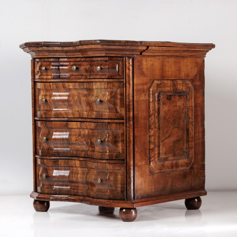 An 18th-century Bavarian walnut chest of drawers featuring a serpentine front, four moulded drawers with brass knobs, panelled sides, and turned bun feet.