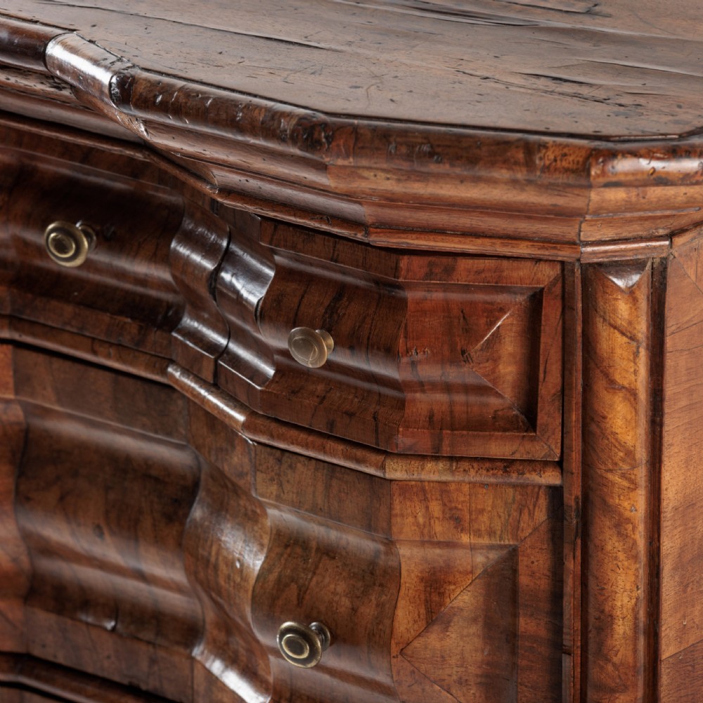 An 18th-century Bavarian walnut chest of drawers featuring a serpentine front, four moulded drawers with brass knobs, panelled sides, and turned bun feet. View V