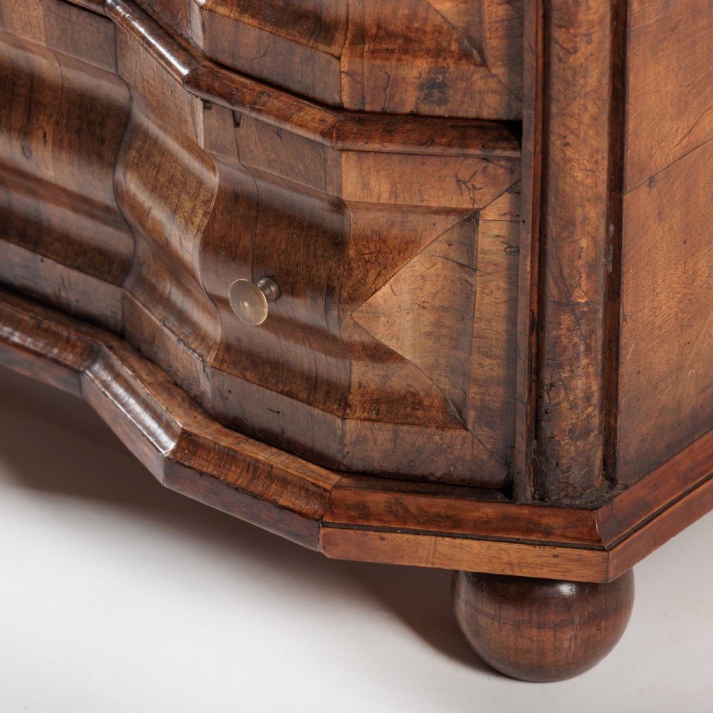 An 18th-century Bavarian walnut chest of drawers featuring a serpentine front, four moulded drawers with brass knobs, panelled sides, and turned bun feet. View VII