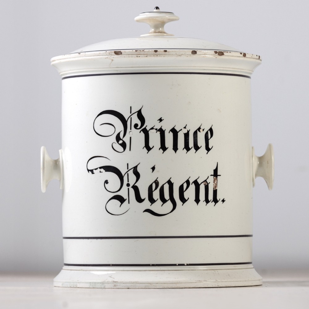 A cylindrical white ceramic jar with a fitted knopped lid, small side handles, and black Gothic script reading Prince Regent.