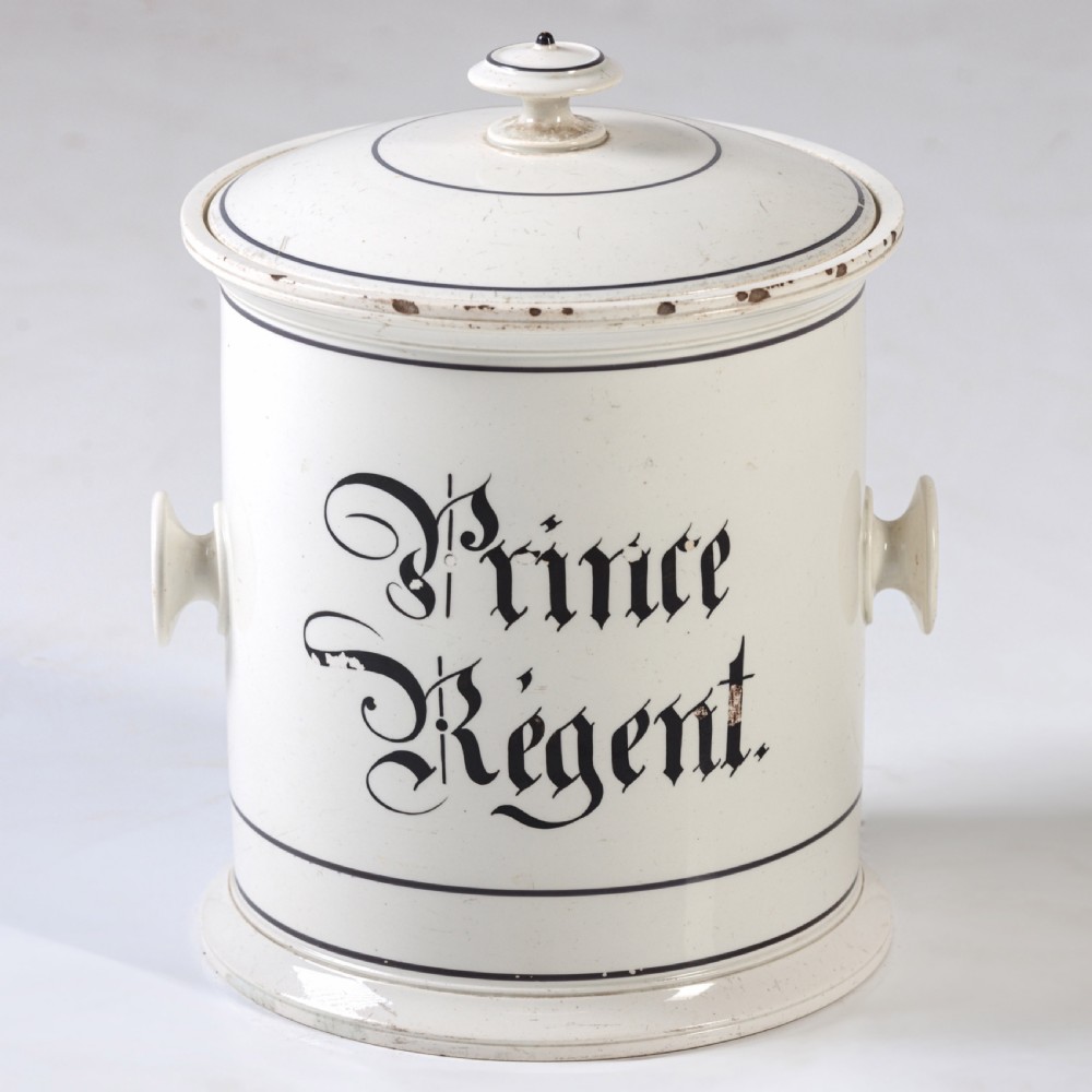 A cylindrical white ceramic jar with a fitted knopped lid, small side handles, and black Gothic script reading Prince Regent. View II
