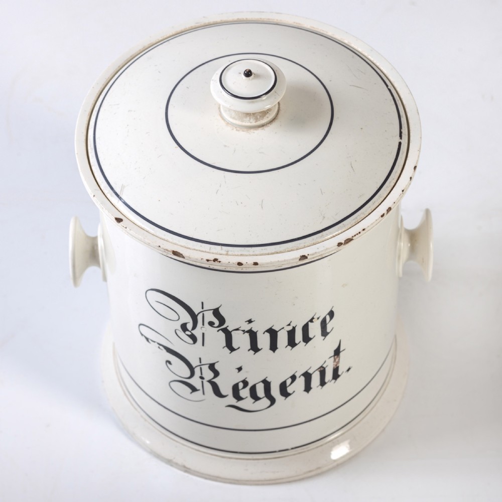 A cylindrical white ceramic jar with a fitted knopped lid, small side handles, and black Gothic script reading Prince Regent. View III