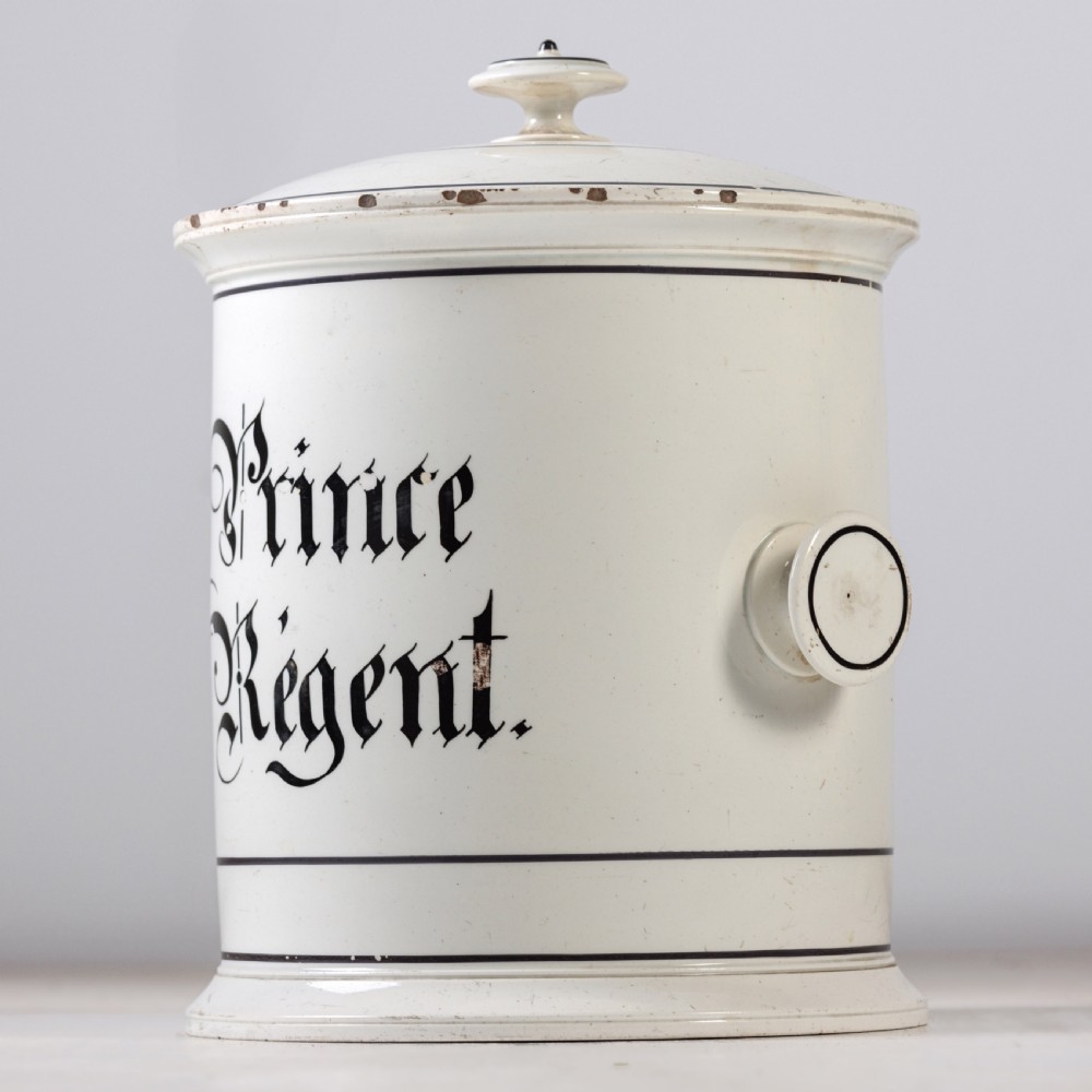A cylindrical white ceramic jar with a fitted knopped lid, small side handles, and black Gothic script reading Prince Regent. View IV
