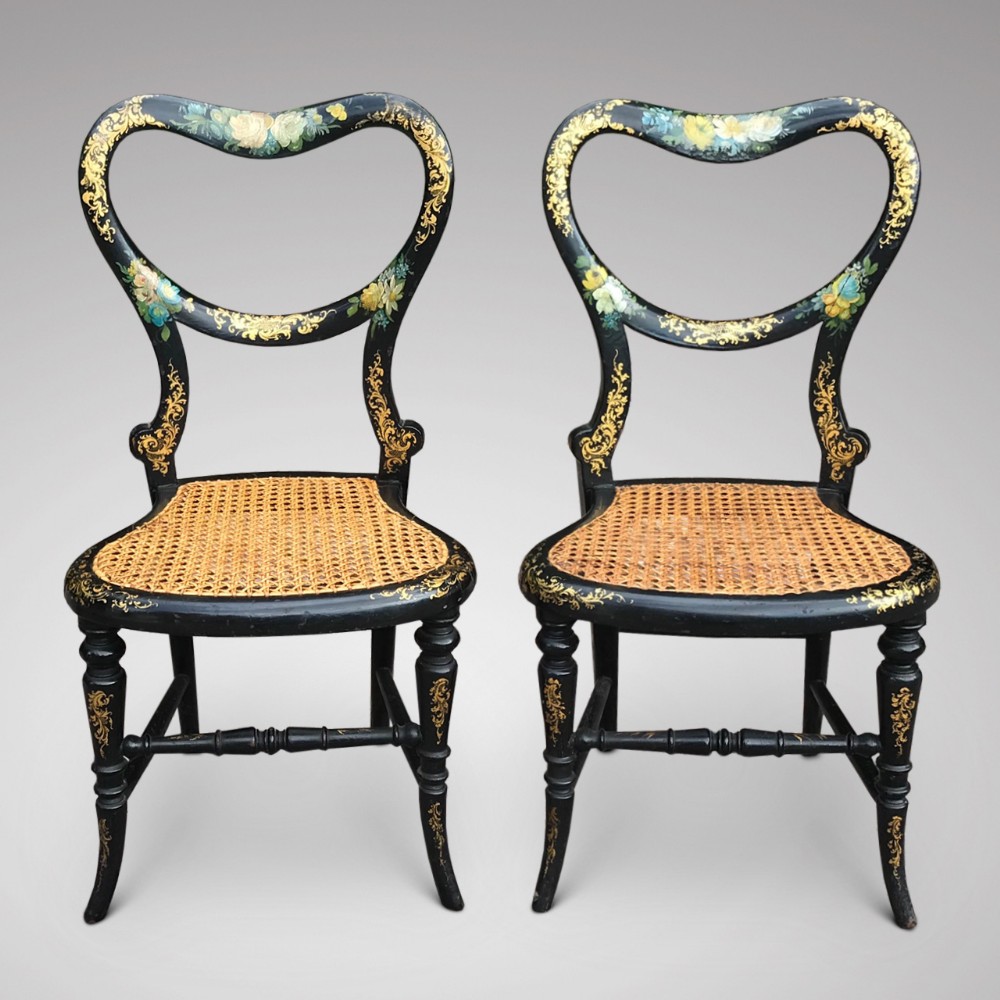 A pair of small Victorian black lacquered papier-mache chairs featuring heart-shaped backrests with floral paintwork, gilt foliage detailing, and woven cane seats.