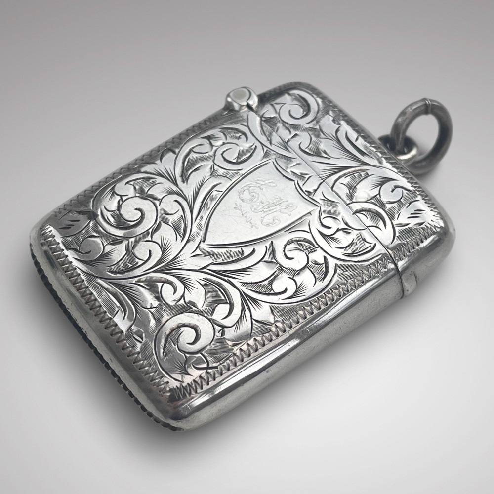 An Edwardian sterling silver pocket vesta case featuring hand-engraved scrollwork, a central monogrammed shield cartouche, and an attached suspension loop.