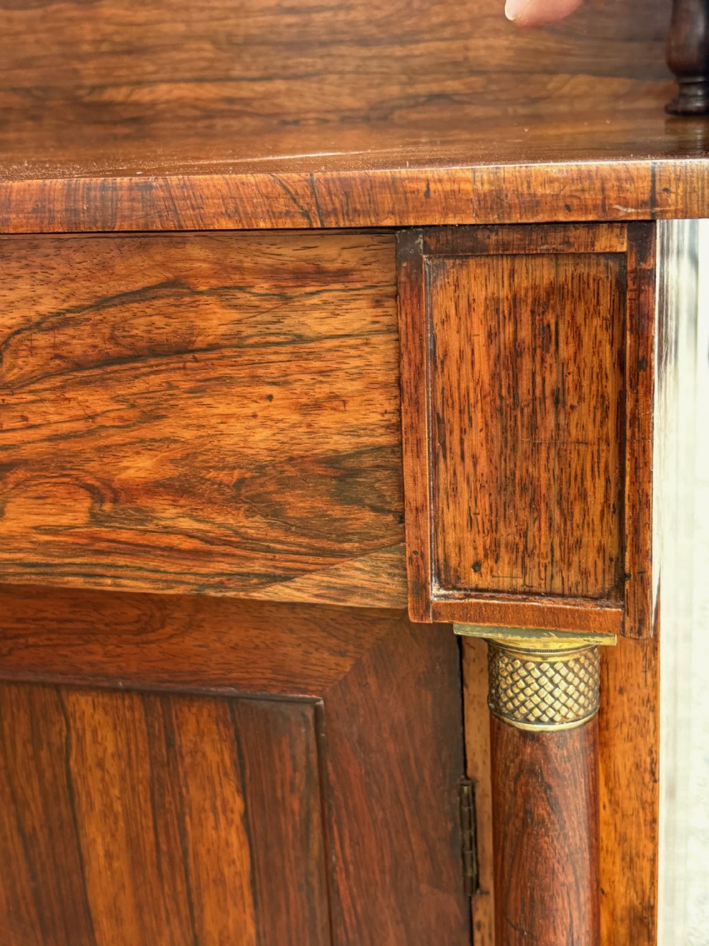 A Regency dark rosewood side cabinet featuring a pedimented upper shelf supported by turned pillars, above a single drawer and a cupboard door flanked by brass-mounted columns. View VIII