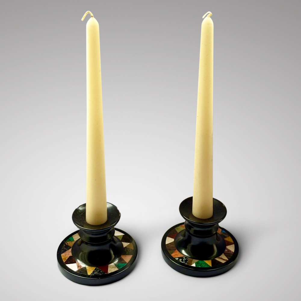 A pair of turned Ashford black marble candlesticks featuring geometric specimen stone inlays around circular bases, fitted with cream taper candles.