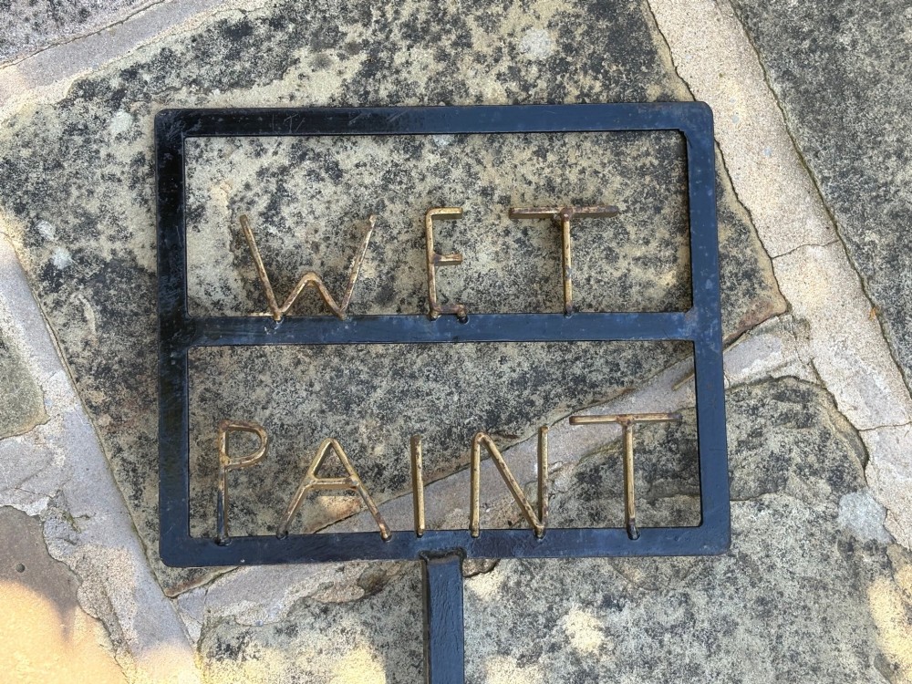 A tall black wrought iron standing sign featuring gold-coloured metal letters spelling WET PAINT framed within a rectangular top section above a scrolled metal base. View III