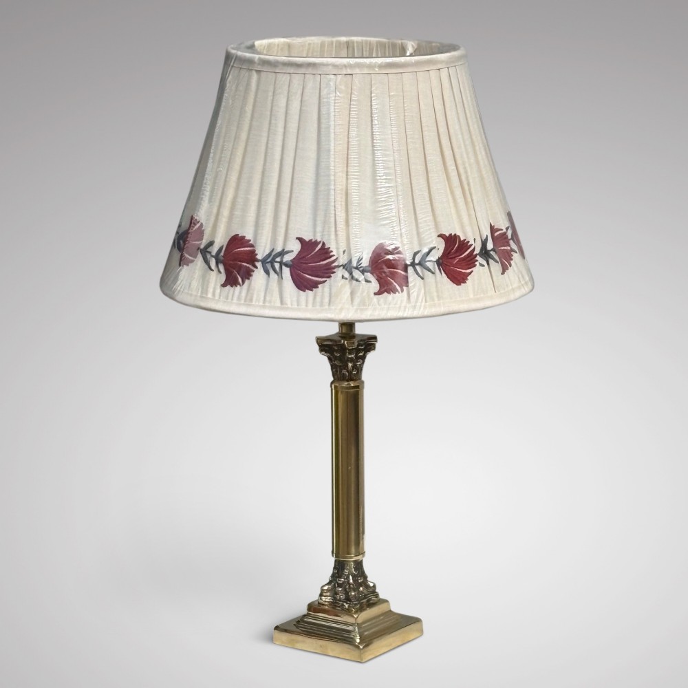 An Edwardian brass table lamp featuring a Corinthian column stem and a pleated cream lampshade decorated with a floral motif.