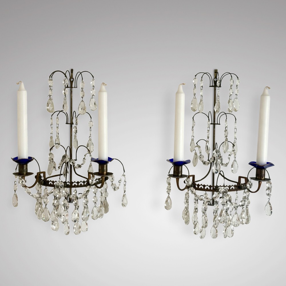 A pair of Swedish antique wall sconces featuring dark brass frames, cascading cut crystal drops, and blue glass drip pans holding white candles.