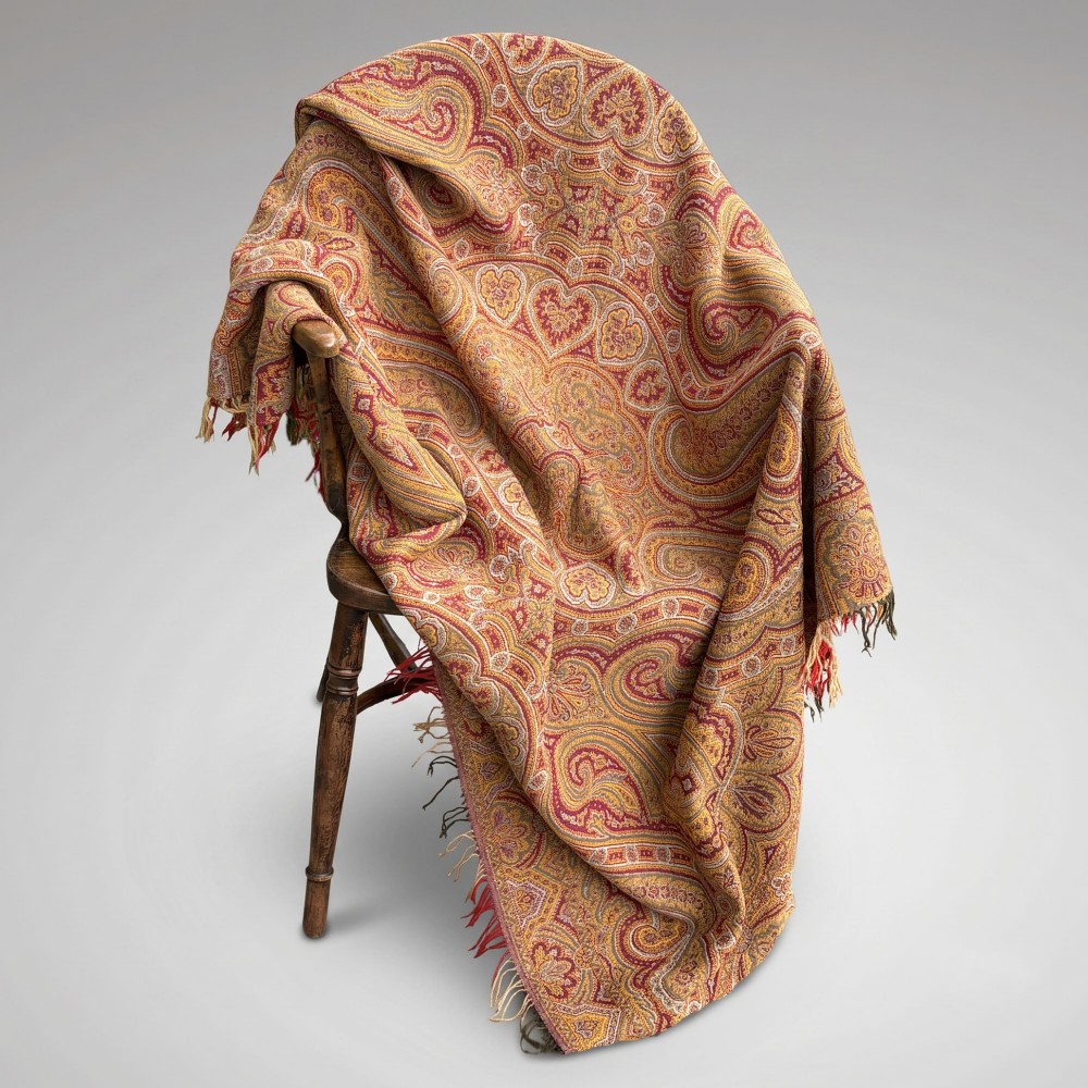 A Victorian wool shawl woven with dense red, ochre, and terracotta paisley motifs, finished with fringed edges.
