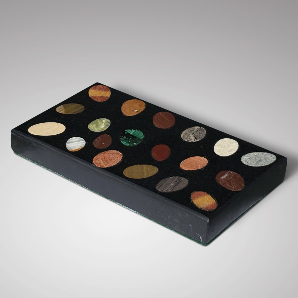 A rectangular black marble paperweight inlaid with twenty oval polished stone specimens of various colours arranged in a grid pattern.