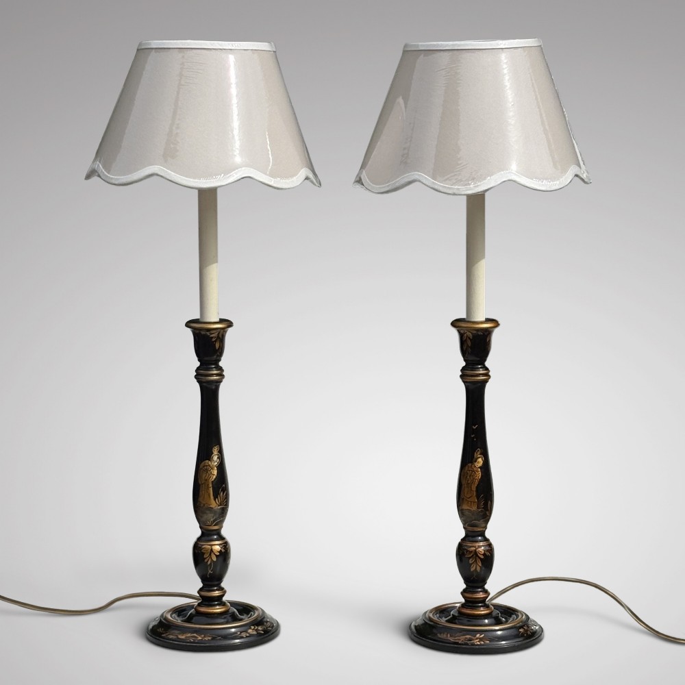 A pair of tall black lacquered table lamps decorated with gold Chinoiserie figures, set on circular stepped bases with cream scalloped fabric shades.