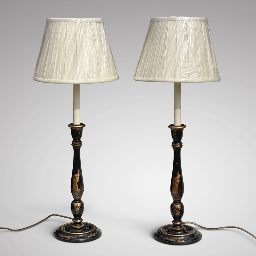 A pair of tall black lacquered table lamps decorated with gold Chinoiserie figures, set on circular stepped bases with cream scalloped fabric shades. View II