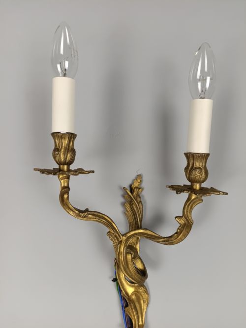 fig 1. A pair of two-arm gilded brass wall sconces featuring ornate Rococo scrolling leaf details and white candle-effect sleeves.