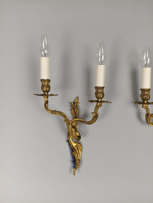 fig 2. A pair of two-arm gilded brass wall sconces featuring ornate Rococo scrolling leaf details and white candle-effect sleeves.