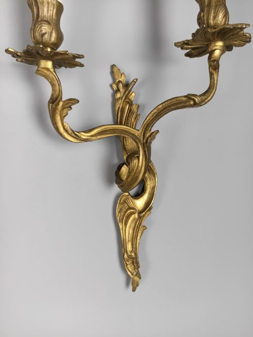 fig 4. A pair of two-arm gilded brass wall sconces featuring ornate Rococo scrolling leaf details and white candle-effect sleeves.
