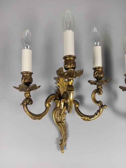 fig 1. A pair of French Rococo style triple-arm gilded bronze wall lights featuring ornate foliage scrollwork and white candle sleeves.