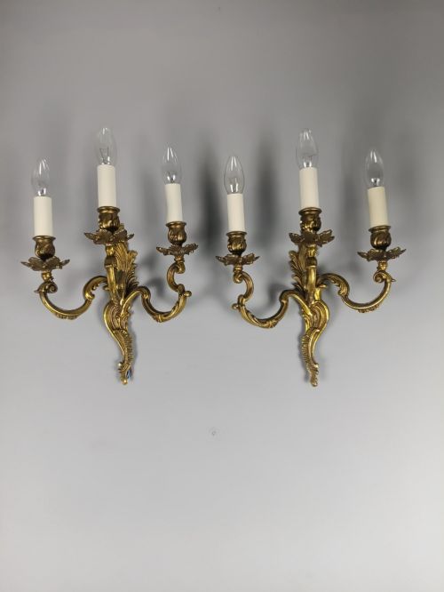 fig 4. A pair of French Rococo style triple-arm gilded bronze wall lights featuring ornate foliage scrollwork and white candle sleeves.