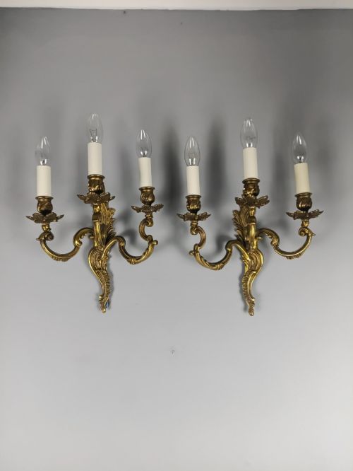 fig 5. A pair of French Rococo style triple-arm gilded bronze wall lights featuring ornate foliage scrollwork and white candle sleeves.