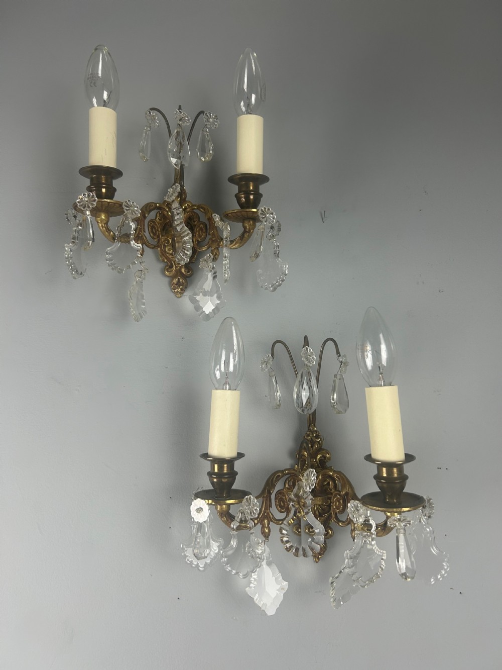 A pair of French gilt brass twin-arm wall lights decorated with scrollwork and hanging crystal drops. View X