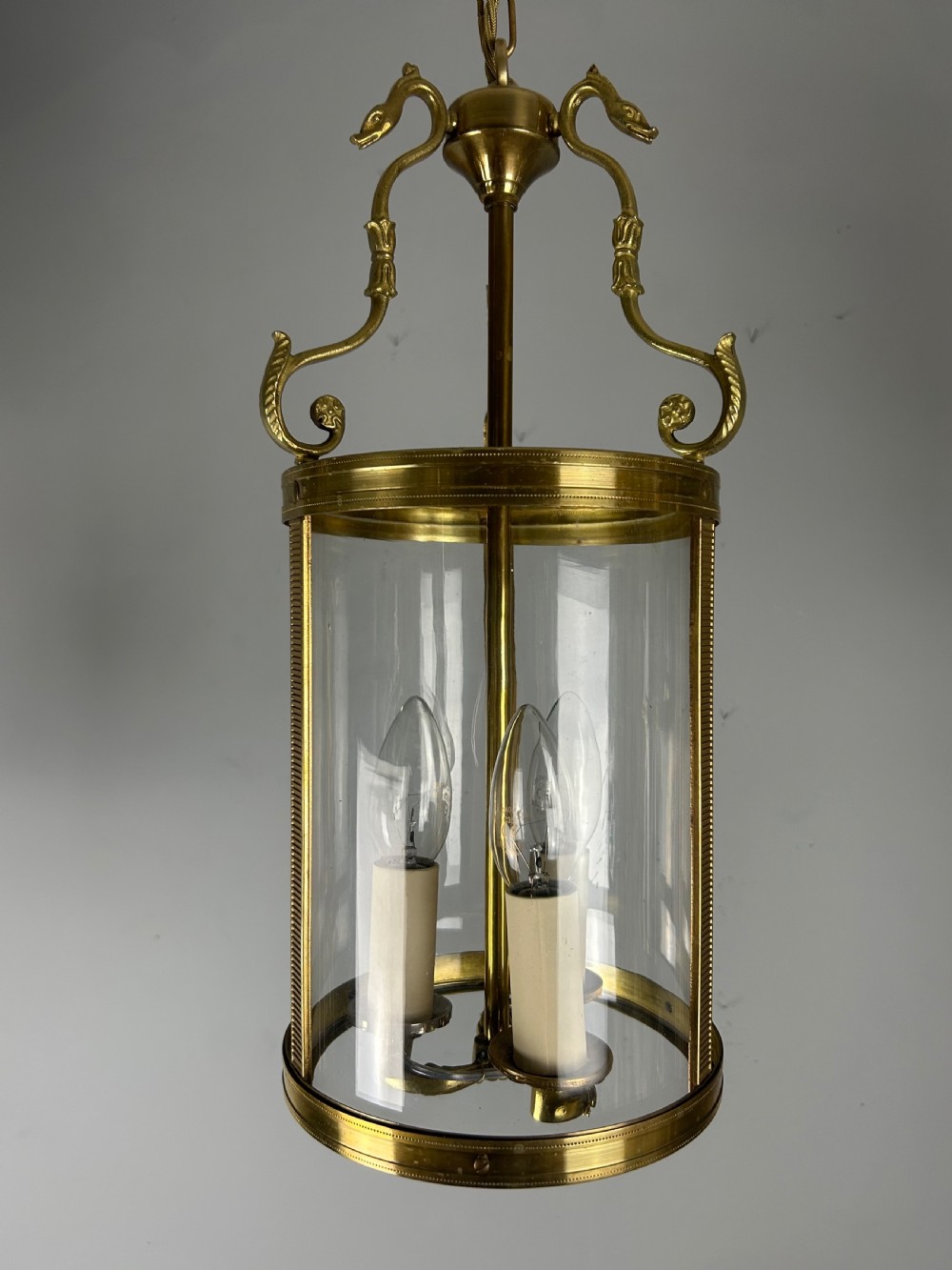 An antique French Empire style hanging brass lantern featuring a clear glass cylinder surrounding three candle-style light sockets and ornate scroll suspension arms.