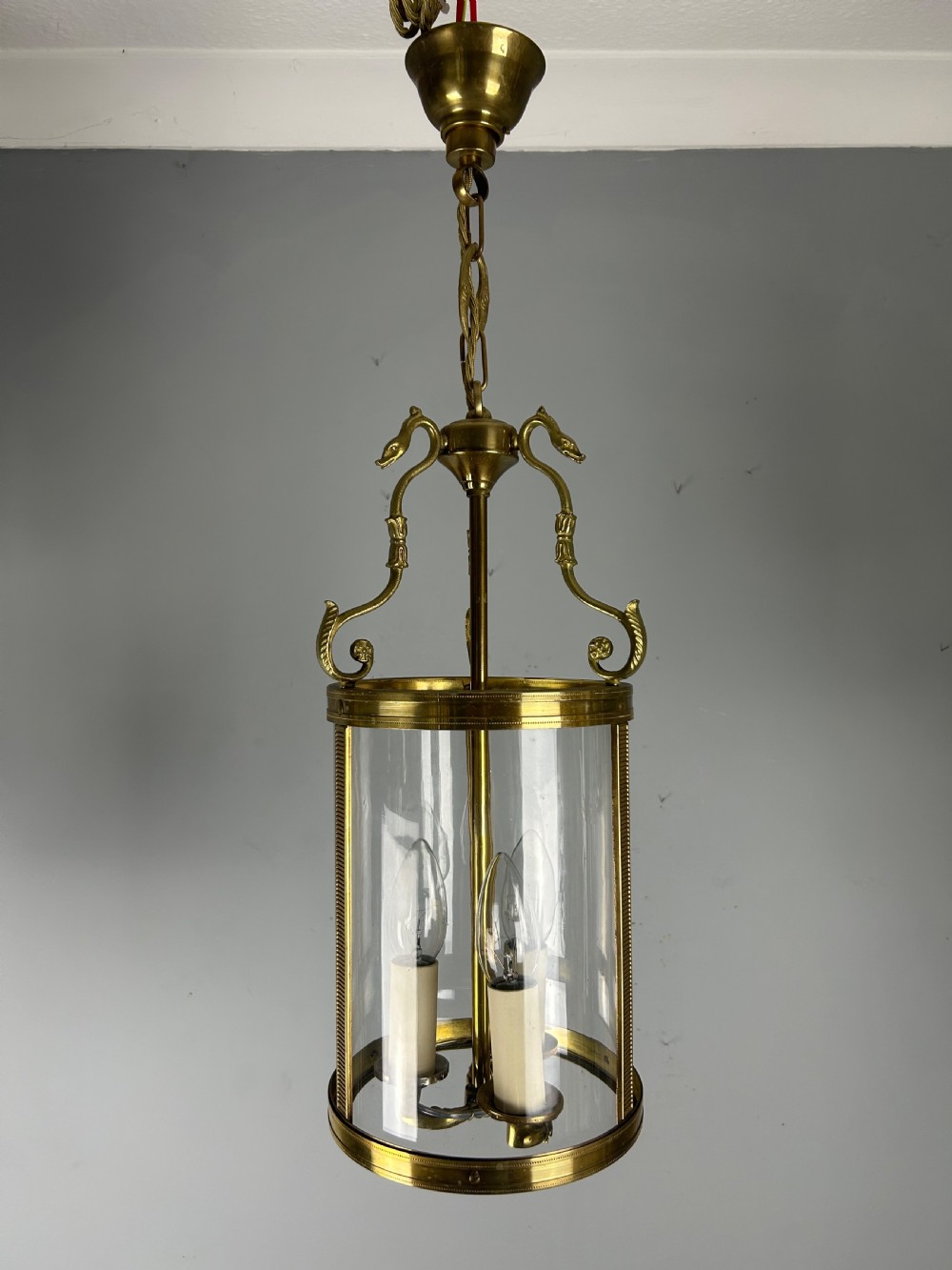 An antique French Empire style hanging brass lantern featuring a clear glass cylinder surrounding three candle-style light sockets and ornate scroll suspension arms. View II