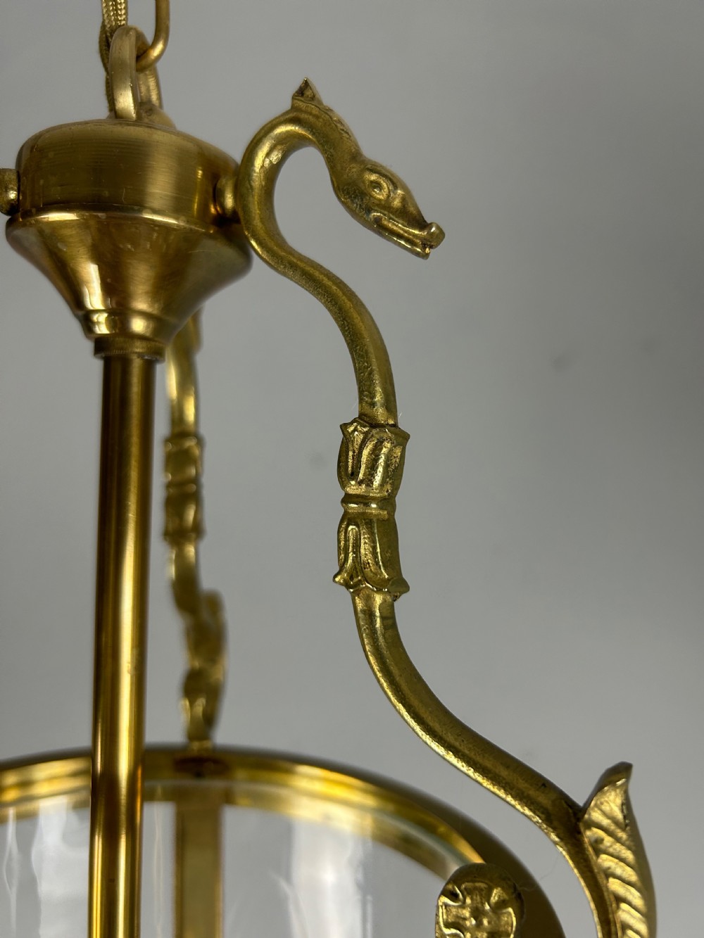 An antique French Empire style hanging brass lantern featuring a clear glass cylinder surrounding three candle-style light sockets and ornate scroll suspension arms. View III